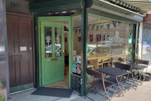 Außensitzbereich des Green Kitchen Vegan Cafe mit Tischen, Stühlen, Pflanzen, einer Matte und einer Glastür mit Postern, die in einen Innenraum mit Tischen, Stühlen und Deckenleuchten führen.