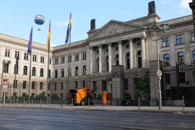 Das Gebäude des Bundestages in Berlin, Deutschland, ein großes Gebäude mit zahlreichen Fenstern, umgeben von Straßenlaternen, Fahrzeugen, Menschen und Bäumen, unter einem bewölkten Himmel.