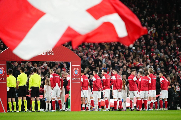 Eine Gruppe von Menschen auf einem Fußballfeld vor einem Bogen mit dem Text "Bayern München vs. Bayern München Wett-Tipps & Vorschau", mit einer roten und weißen Flagge im Vordergrund und einer großen Menge im Stadionhintergrund.
