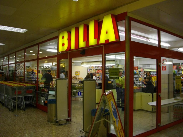 Ein Supermarkt mit einem Eingang in der Mitte, Einkaufswagen aufgereiht auf der linken Seite und einigen Einkäufern, die unter Deckenbeleuchtung Produkte betrachten.