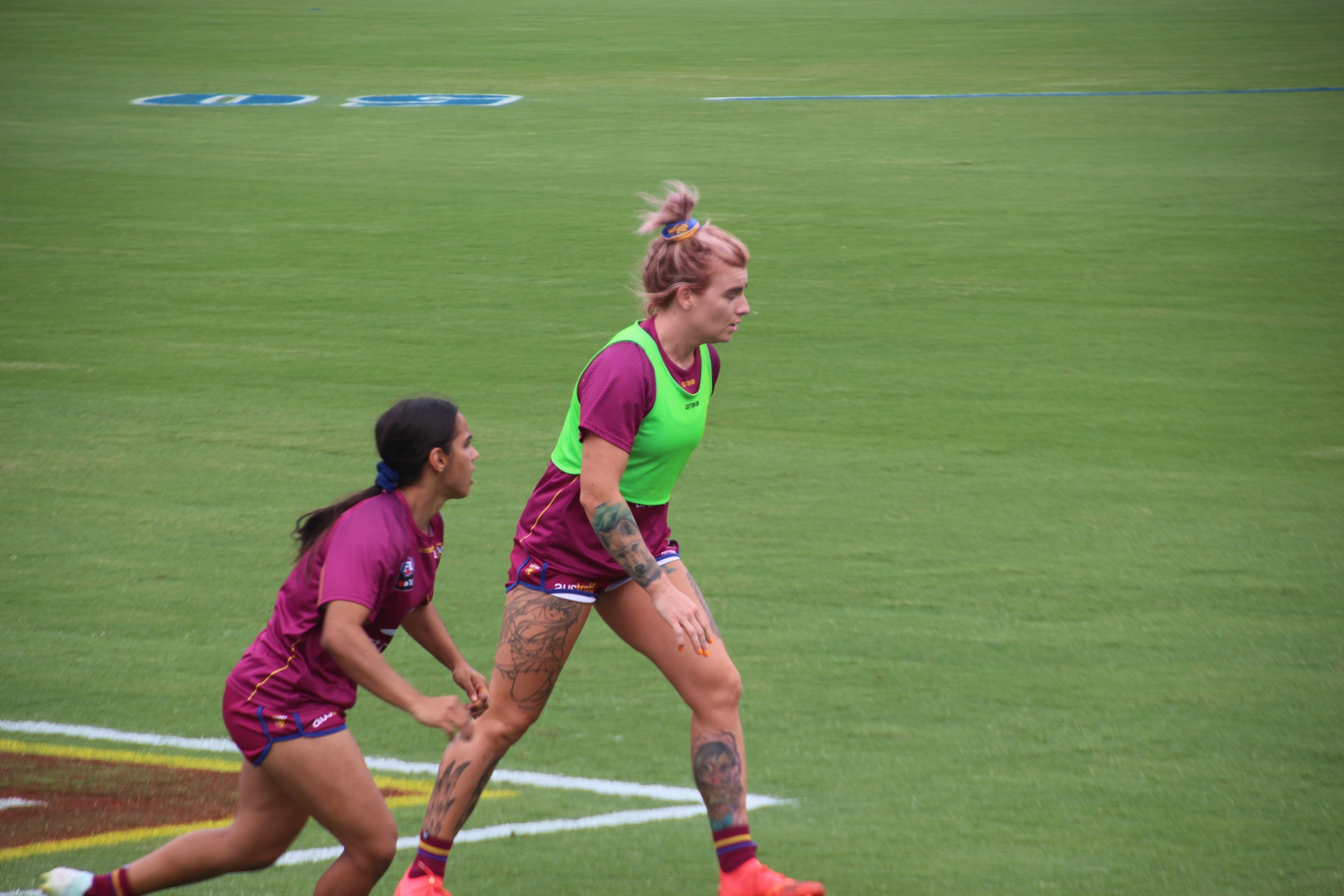 Zwei Frauen in Sportkleidung spielen Fußball, eine in einem grünen T-Shirt und Shorts, die andere in einem pinken Kleid, beide mit sichtbaren Tattoos an Händen und Füßen.