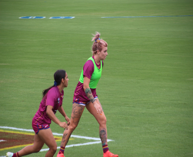 Zwei Frauen in Sportkleidung spielen Fußball, eine in einem grünen T-Shirt und Shorts, die andere in einem pinken Kleid, beide mit sichtbaren Tattoos an Händen und Füßen.