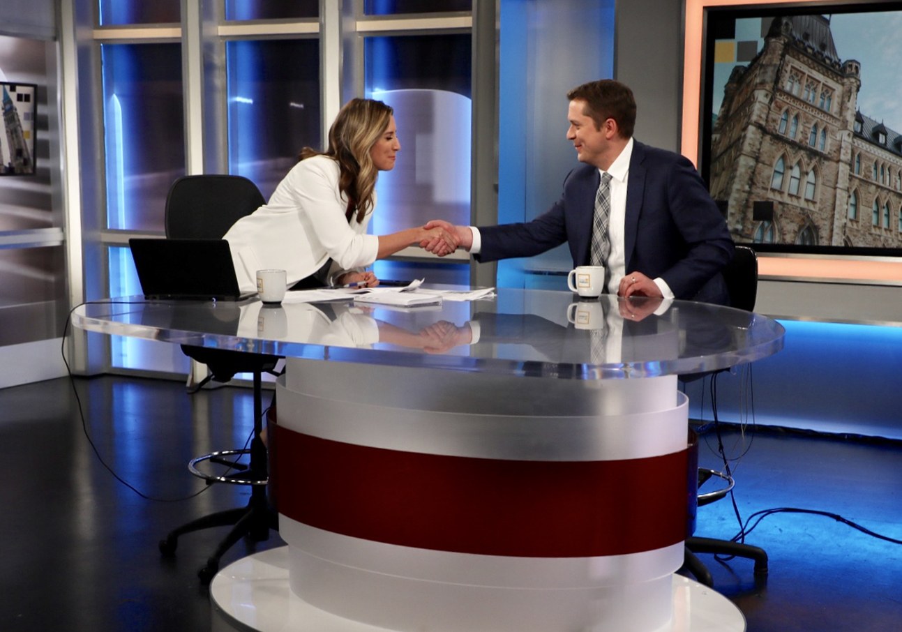 Zwei Personen, die vor einem Fernseher Hände schütteln, mit einem Tisch dazwischen, auf dem Tassen, Papiere, ein Laptop und andere Gegenstände stehen, sowie Glasfenster und ein wandmontierter Bildschirm im Hintergrund zu sehen sind.