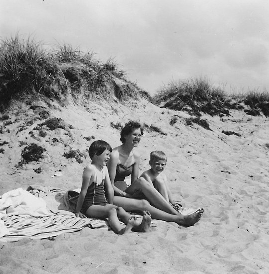 Schwarzes und weißes Foto einer Frau in einem Bikini und zwei Kindern in Badebekleidung, die auf einer Decke auf einem sandigen Strand mit Gras und Himmel im Hintergrund sitzen.