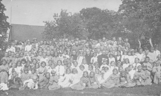 Schwarz-weiß-Foto einer Gruppe von Kindern, die vor einem Haus mit Bäumen und einem klaren Himmel posieren.