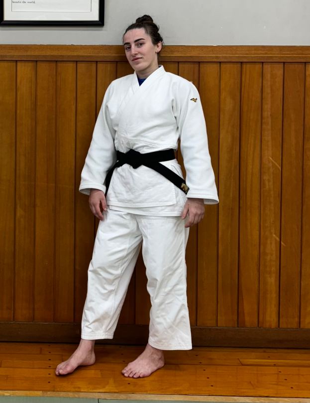 Eine Frau in einem weißen Judo-Anzug steht selbstbewusst vor einer hölzernen Wand mit einem Rahmen, der eine entschlossene Miene zeigt.