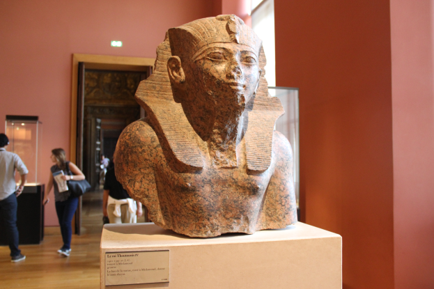 Ein ägyptischer Pharaonenkopf wird in einem Museum ausgestellt, mit einer Texttafel, einer Gruppe von Menschen, einer Frau mit Tasche, einer Tür, einer Wand und einer Decke mit Leuchten.