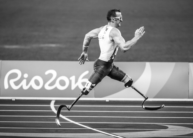 Ein Mann mit einer Prothese läuft auf einer Laufbahn mit einem 'Rio 2016 Paralympische Spiele'-Schild im Hintergrund, dargestellt in Schwarz-Weiß.