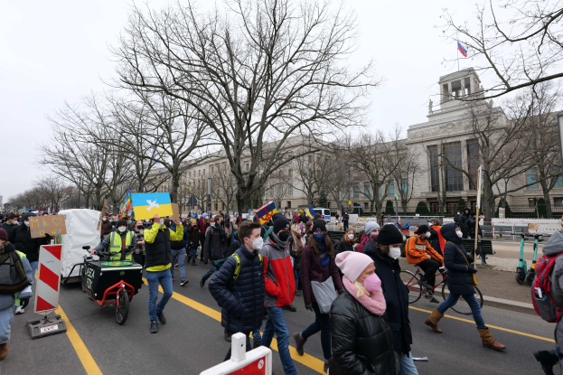 Eine große Gruppe von Menschen nimmt an einer Protestmarsch in Washington, D.C. am 21. Januar 2020 teil, geht eine Straße entlang und hält Plakate und Schilder in die Höhe, während andere Fahrräder fahren, mit Schilderständen, Bäumen und einem klaren blauen Himmel im Hintergrund.