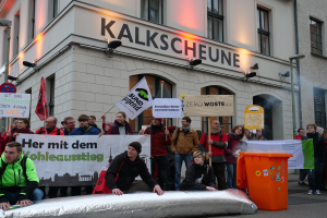 Eine Gruppe von Menschen mit Schildern und Plakaten steht vor einem Gebäude, mit zwei Personen im Vordergrund und einem Mülleimer rechts, während einer Protestaktion in Deutschland.