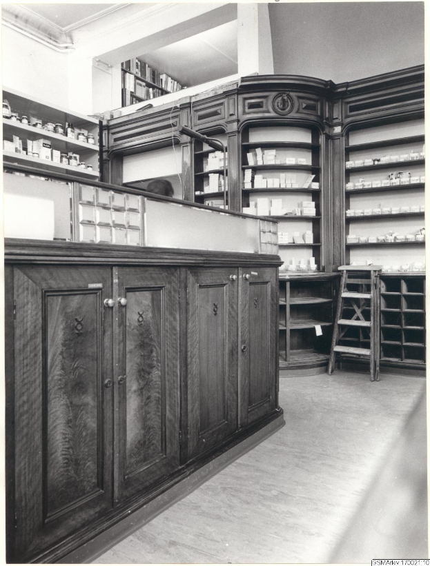 Ein Schwarz-Weiß-Foto eines Apothekeninneren mit einer Theke, Schränken, einer Leiter und Regalen, die mit verschiedenen Gegenständen gefüllt sind, mit Text oben drauf.