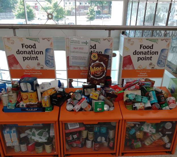 Orange Schränke gefüllt mit Lebensmittelspenden, darunter Flaschen, Dosen und Kartons, mit informativen Schildern; draußen vor den Glasfenstern sind Fahrzeuge, Bäume, Gebäude und Himmel zu sehen.