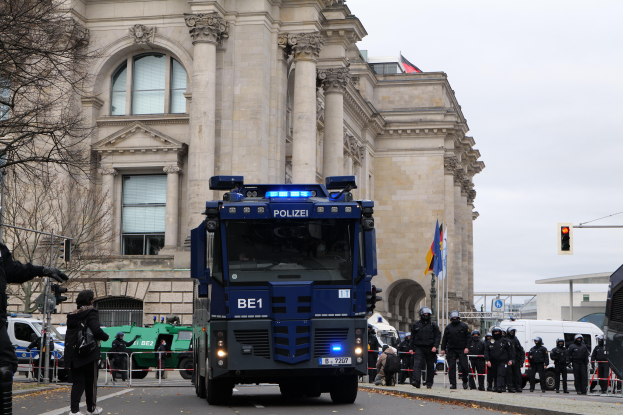 Eine Gruppe von Polizisten steht vor einem großen Gebäude mit Fenstern, Säulen und Bogengängen, mit Fahrzeugen auf der Straße und einer Person mit einer Kamera auf der linken Seite, unter einem klaren blauen Himmel mit Bäumen, Ampeln und Flaggen im Hintergrund.