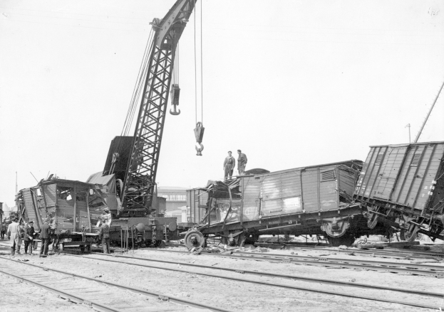Schwarzes und weißes Bild von einer Zugkatastrophe mit einem Kran, der einen Zugwagen auf die Schienen hebt, mehreren Menschen in der Nähe und Gebäuden im Hintergrund bei klarem Himmel.