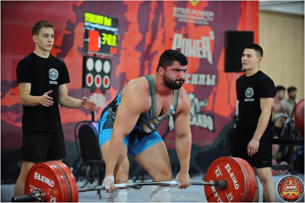 Ein Mann, der eine Hantel in einem Gewichtheben-Wettbewerb hochhebt, umgeben von Zuschauern, mit Wettkampfausrüstung und Beschilderung, die die 2018 World Weightlifting Championships anzeigt.
