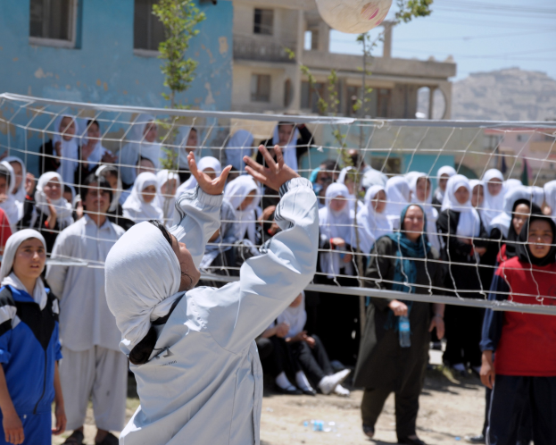 Kinder spielen Volleyball mit einem Netz, vor einer Menge, mit Gebäuden, Bäumen, Höllen und einem klaren blauen Himmel im Hintergrund.