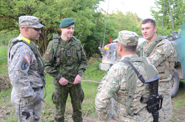 Vier Armee-Männer in Uniform und Mützen stehen und sprechen, einer hält eine Waffe, ein Fahrzeug auf der rechten Seite und Bäume im Hintergrund.