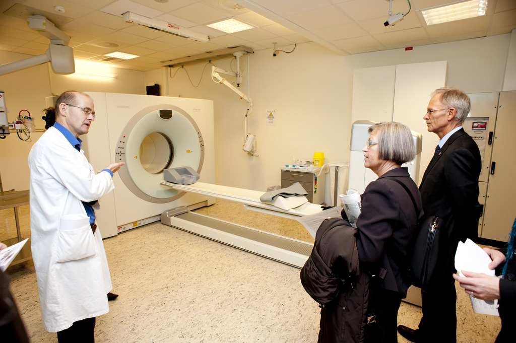 Ein Arzt in einem weißen Kittel erklärt eine CT-Scan-Maschine zwei Personen in schwarzen Mänteln, die aufmerksam zuhören.