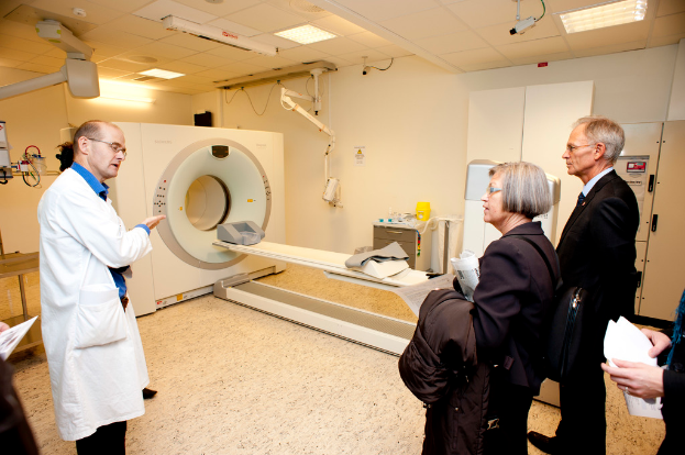 Ein Arzt in einem weißen Kittel erklärt eine CT-Scan-Maschine zwei Personen in schwarzen Mänteln, die aufmerksam zuhören.