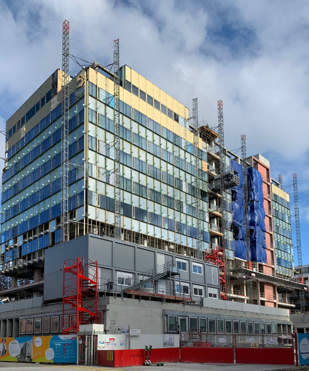 Großes Gebäude im Bau in einer Stadt mit Glasfenstern, Metallrahmen, einer Treppe, einem Zaun, einem Schild, Polen, einem Straßenpfahl, Bäumen und einem bewölkten Himmel.