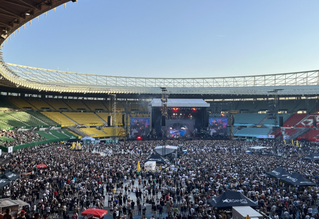 Eine große Menschenmenge versammelt sich vor einem Stadion mit Schirmen und Zelten und wartet auf ein Musikfestival, während Bühnenlichter und Bildschirme im Hintergrund zu sehen sind.