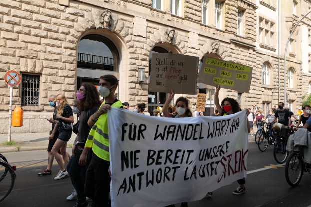 Eine Gruppe von Menschen marschiert bei einer Demonstration auf einer Stadtstraße, einige halten Schilder und andere fahren Fahrräder, im Hintergrund ein historisches Gebäude mit Rundbogenfenstern, Säulen und Skulpturen.