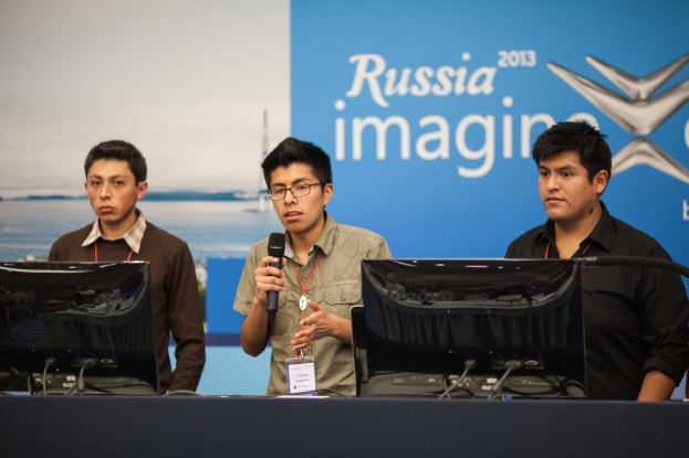 Drei Jungen stehen neben einem Schreibtisch mit zwei Computern, einer spricht in ein Mikrofon, tragen ID-Karten mit roten Markierungen, vor einer Wand mit einer Werbetafel, auf der "Russia Imagine 2013" steht.