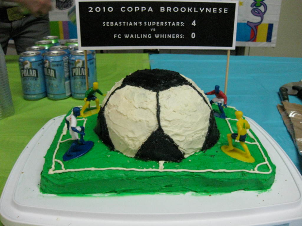 Ein kuchenförmiger Fußballstadion-Kuchen mit einem Fußball und vier Spielern, umgeben von Softdrink-Dosen, mit einem Banner im Hintergrund und einer Person auf der linken oberen Seite, mit einer Tafel auf dem Kuchen.