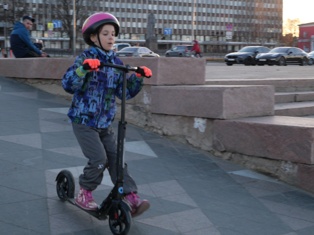 Ein junger Junge mit Helm und Handschuhen fährt auf einem Roller eine Treppe auf dem Gehweg hinunter, mit Autos, Menschen, Bäumen, Pfosten, Brettern, Gebäuden und einem klaren blauen Himmel im Hintergrund.