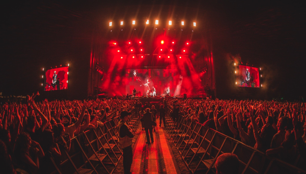 Eine große Menschenmenge steht vor einer hell erleuchteten Bühne mit Geländern, auf der Musiker spielen und andere zuschauen, vor einem dunklen Hintergrund.