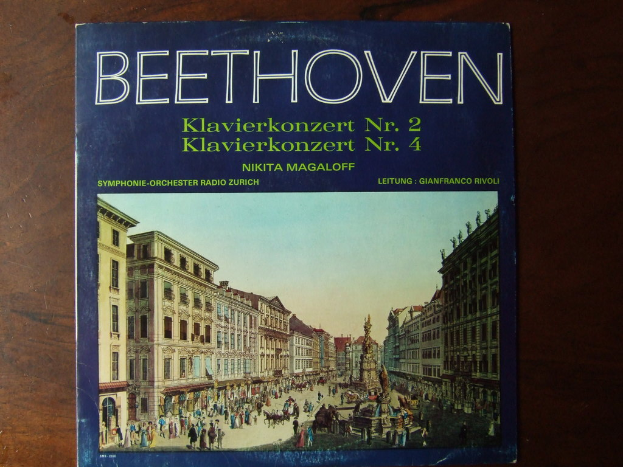 Ein Buch auf einer hölzernen Oberfläche, das eine Szene mit vielen Menschen auf einer Straße, Gebäuden auf beiden Seiten, einem Himmel darüber und einer Skulptur in der Mitte zeigt.
