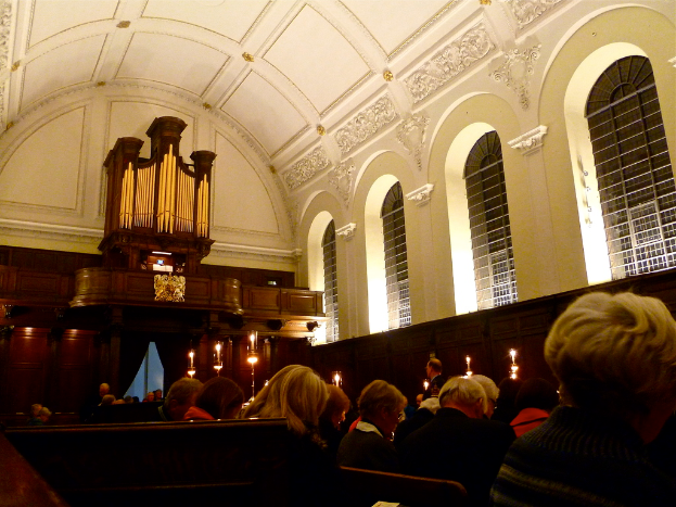 Das Innere einer Kirche mit Männern und Frauen, die sitzen, Kerzen vor ihnen und weißen Wänden und Dach.