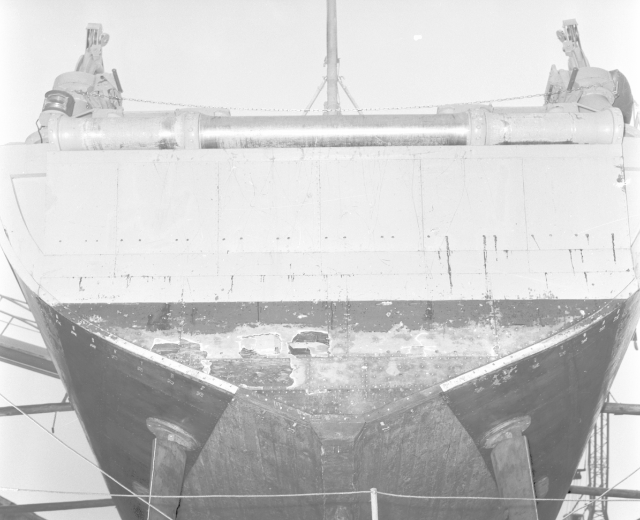 Ein Schwarz-Weiß-Foto eines großen, verrosteten Schiffs in einem Trockendock, umgeben von Pfählen und Seilen.