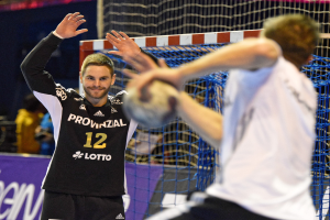 Zwei Männer spielen Handball vor einem Tor mit Netz und einem Banner mit Text im Hintergrund und einigen Menschen in einer Arena.