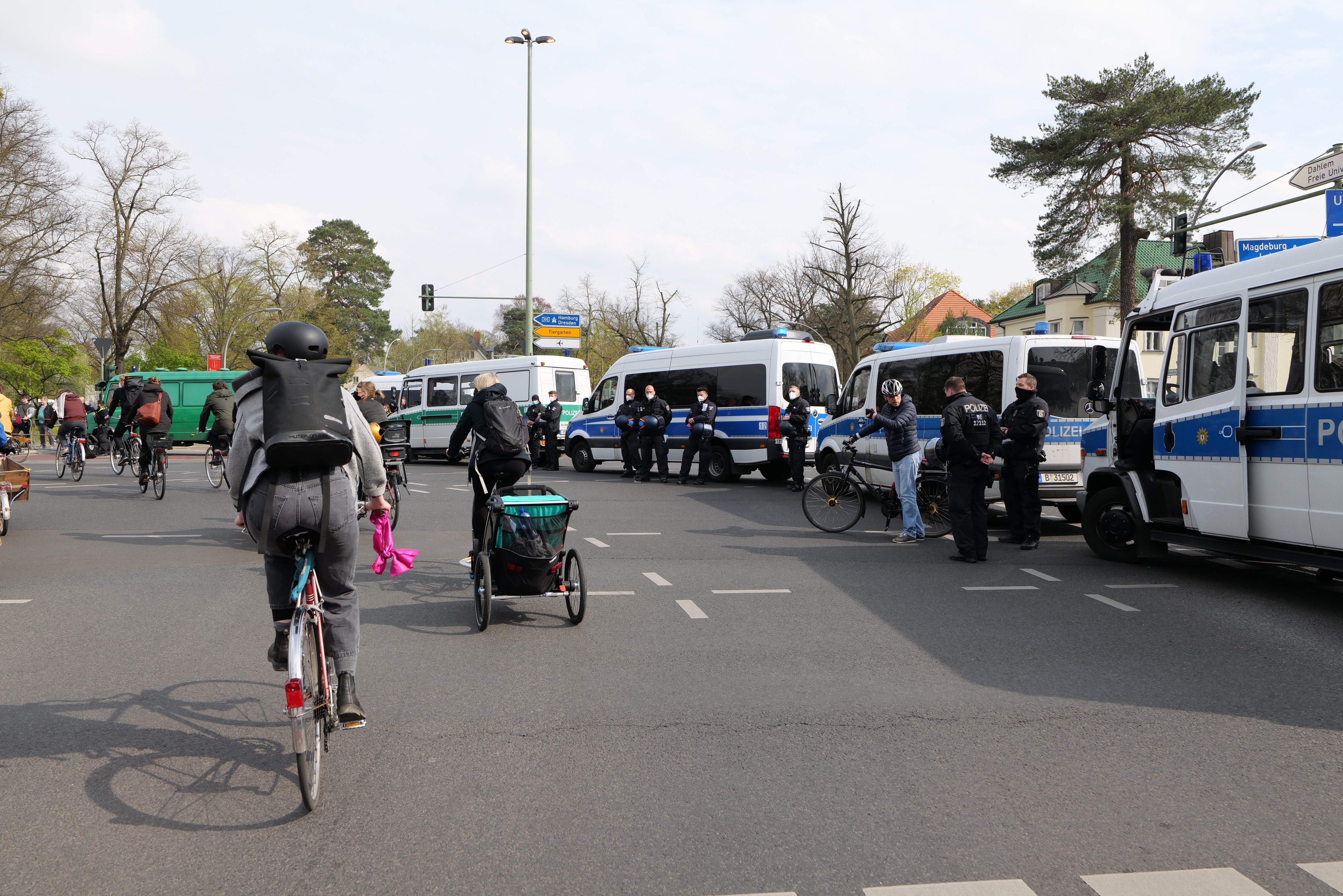 Eine Gruppe von Menschen, die auf Fahrrädern eine Straße entlangfahren, mit Polizeiwagen und anderen Fahrzeugen, einige tragen Helme und haben Taschen oder schieben Kinderwagen, mit Bäumen, Gebäuden und Verkehrszeichen im Hintergrund.