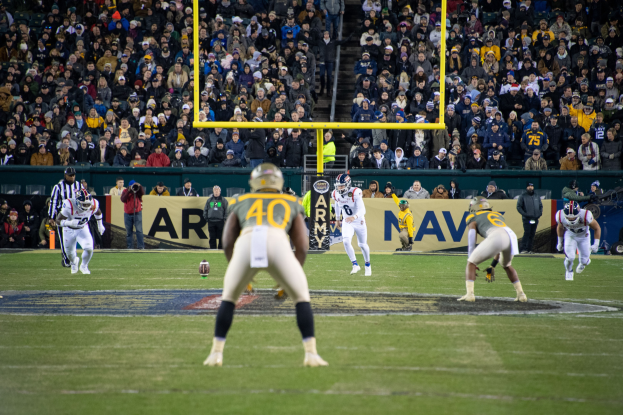 Fußballspieler in Sportkleidung und Helmen konkurrieren auf einem Feld mit Zuschauern im Hintergrund, Stadionstrukturen und sichtbare Beschriftung.