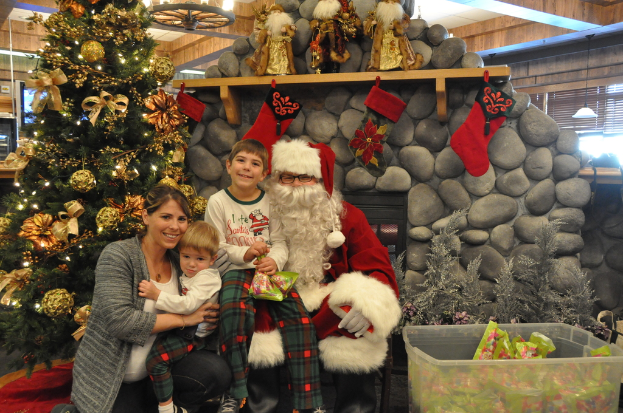Eine Frau, Kinder und ein als Santa Claus verkleideter Mann mit Brille und Handschuhen stehen neben einem geschmückten Weihnachtsbaum, einer Wand mit Steinen, Spielzeugen, Dekorationen und einem Korb mit Gegenständen, mit Lichtern im Hintergrund.