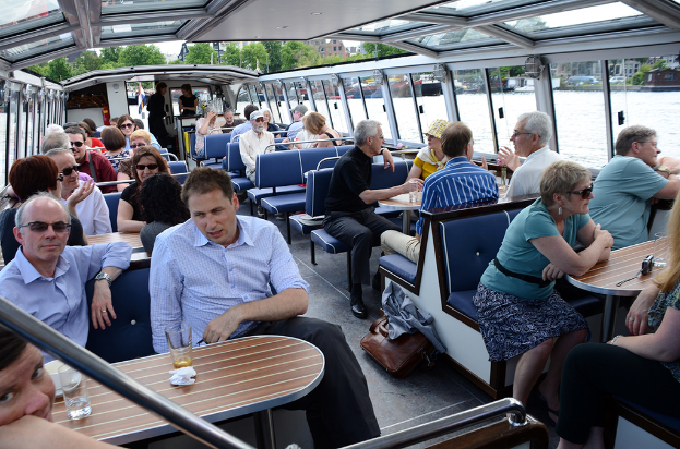 Ein Boot-Innenraum mit Sitzreihen, die von Menschen besetzt sind, Tischen mit Gläsern und Schüsseln davor, einer Tasche auf dem Boden, Wasser im Hintergrund, Gebäuden auf der rechten Seite und kleinen Pflanzen über dem Boot.