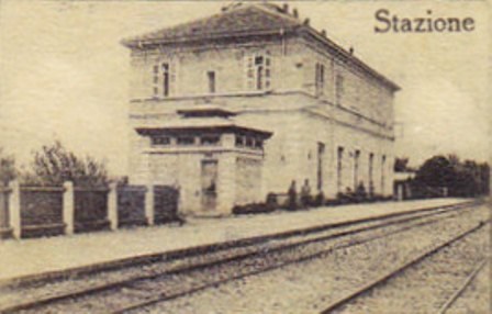 Altes Schwarz-Weiß-Foto eines Bahnhofs mit einem zentralen Gebäude, Bäumen, einem Zaun auf der linken Seite, Bahnschienen unten und Text oben.