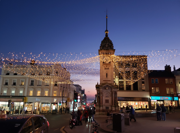 Eine belebte Stadtstraße bei Nacht mit Menschen, Fahrzeugen, Fahrrädern, Mülltonnen, Gebäuden, Laternenmasten, Verkehrsampeln und einem Uhrenturm im Hintergrund, beleuchtet von Weihnachtslichtern.