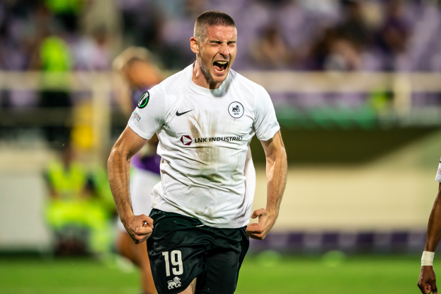Ein Mann in weißem Hemd und schwarzen Shorts rennt auf einem Fußballfeld, feiert ein Tor und ist von einer Gruppe Menschen umgeben, mit einem unscharfen Hintergrund.