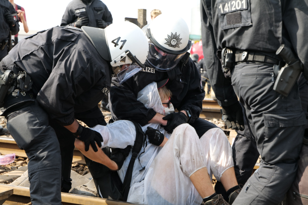 Eine Gruppe von Polizisten in Einsatzausrüstung nimmt einen Mann in einem weißen Kleid auf einem Gleis fest, mit anderen im Hintergrund.