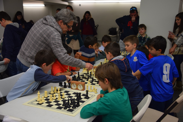 Kinder spielen Schach in einem Wettbewerb, während Erwachsene im Hintergrund und an den Seiten zuschauen.