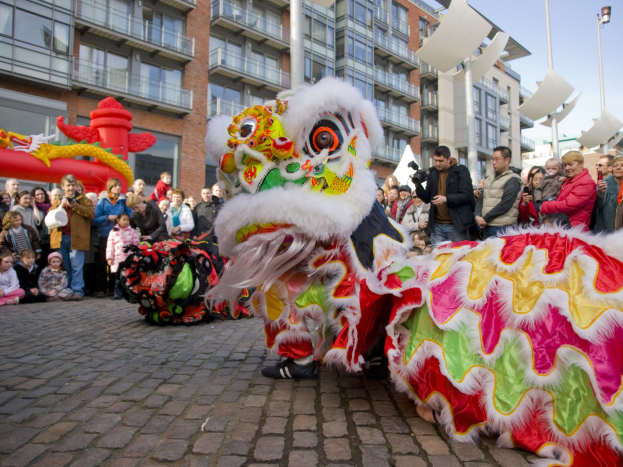 Ein farbenfrohes chinesisches Neujahrsfest in Amsterdam mit einem Löwen tanzen im Vordergrund, umgeben von einer Menschenmenge und Gebäuden im Hintergrund unter einem klaren blauen Himmel.