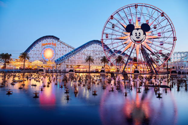 Eine lebendige Disneyland California Adventure-Szene bei Dämmerung mit einem riesigen Mickey-Maus-Rad im Vordergrund, einer Achterbahn im Hintergrund und Booten auf dem Wasser sowie einem Himmel aus Orangen, Pink- und Purpurtönen und silhouettenartigen Bäumen.