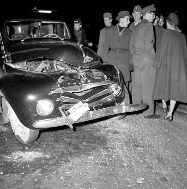 Ein Schwarz-Weiß-Bild eines verbeulten Autos am Straßenrand mit einer Gruppe von Menschen drumherum, die den erheblichen Schaden am Fahrzeug begutachten.