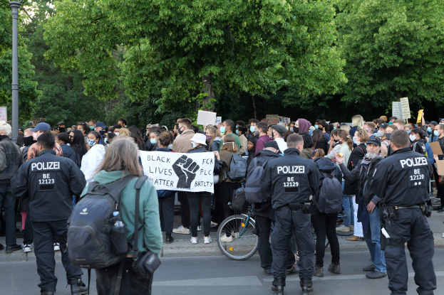 Eine große Gruppe von Menschen steht an der Straße, einige halten Schilder und andere tragen Kappen und Taschen, mit einem Fahrrad im Vordergrund und Bäumen und einem Pfahl im Hintergrund während einer Black Lives Matter Demonstration in Berlin.