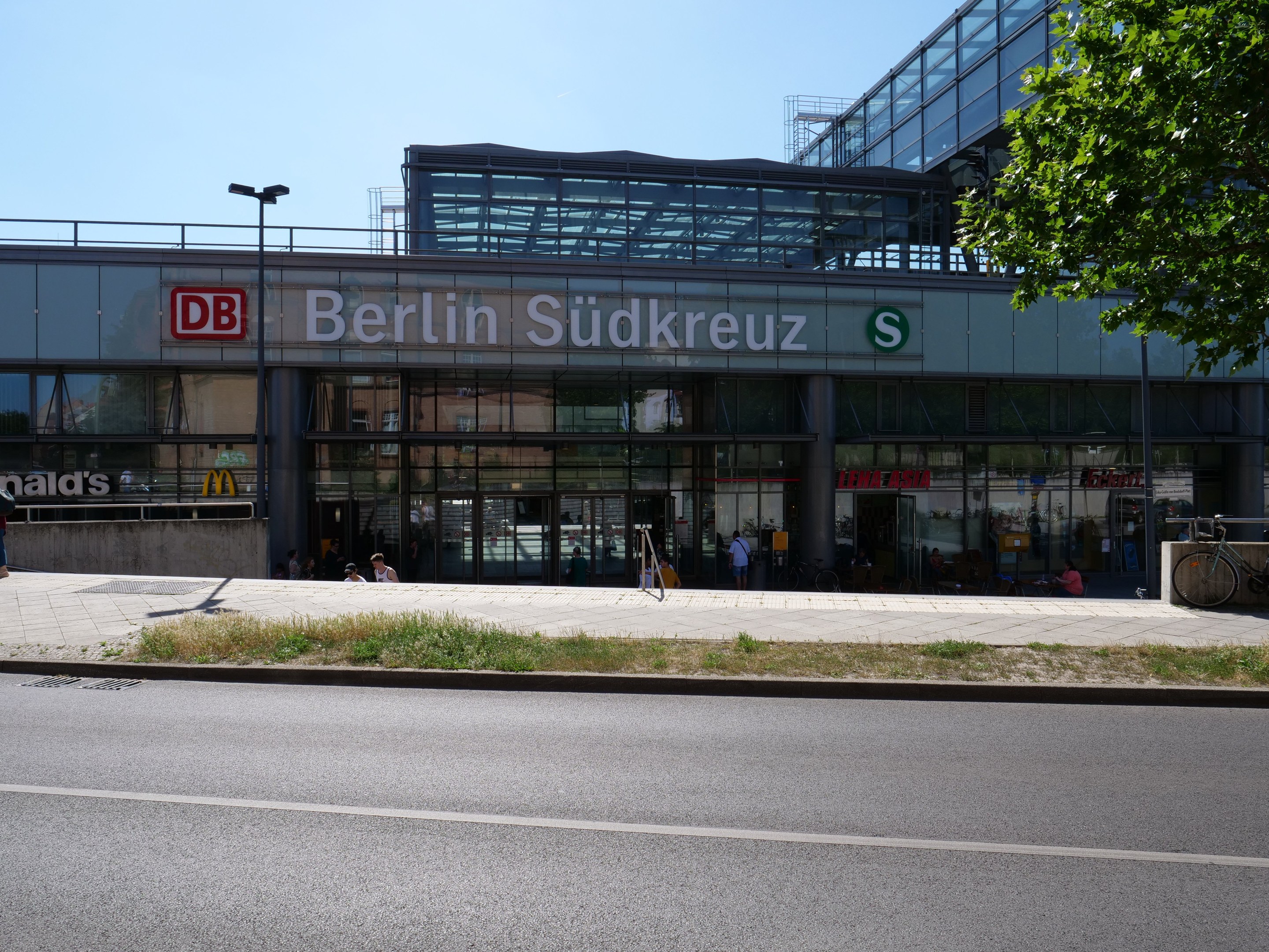 Großes gläsernes Berliner Südkreuz-Bahnhof mit sichtbarem Text, umgeben von Straßenmöbeln, Fahrzeugen, Fußgängern, Fahrrädern, Bäumen und einem klaren Himmel.