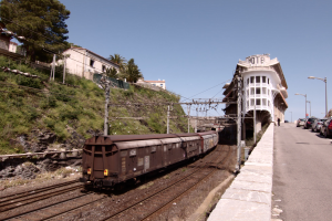 Güterzug fährt auf Gleisen neben einem Gebäude mit parkenden Autos auf einem Bahnsteig auf der rechten Seite, vor Gebäuden, Bäumen, Strommasten, Drähten und einem klaren blauen Himmel.
