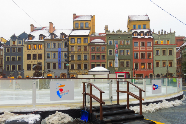 Eislaufbahn in einem Stadtplatz umgeben von Geb├Ąuden, Treppen, Gel├Ąndern, Informationsschildern, schneebedecktem Boden, Laternenmasten, B├Ąumen und einem Himmel.